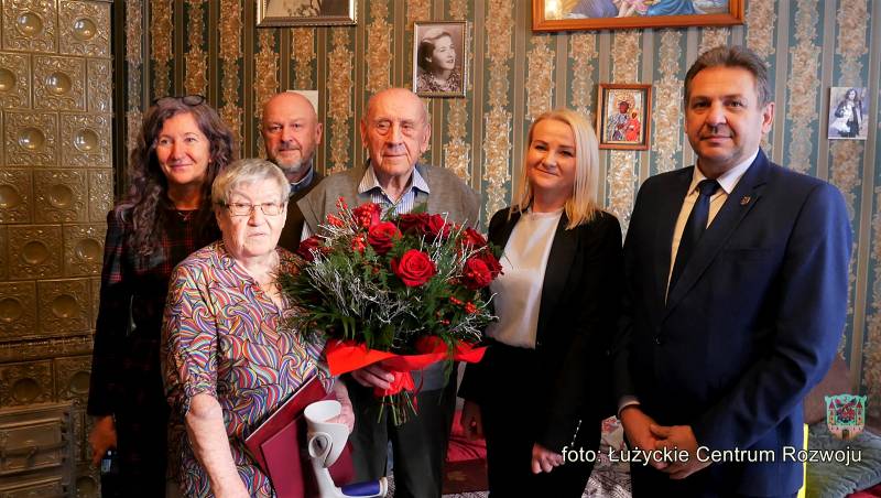 Zdjęcie grupowe - Jubliaci, burmistrz Lubania, kierowniczka USC oraz córka i zięć Jubilatów stoją na tle ściany, na której wiszą fotografie rodzinne